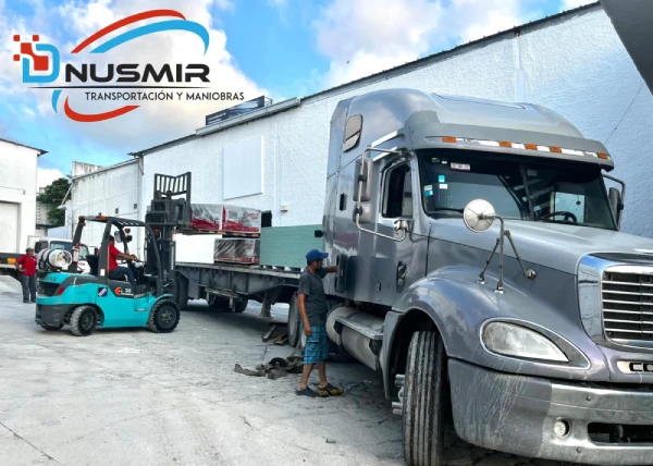 Montacargas cargando mercancía en plataforma de tráiler junto a camión gris en patio industrial