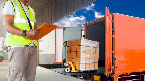 Operario con chaleco reflectante revisa la carga paletizada mientras se introduce en camión de caja con puertas abiertas