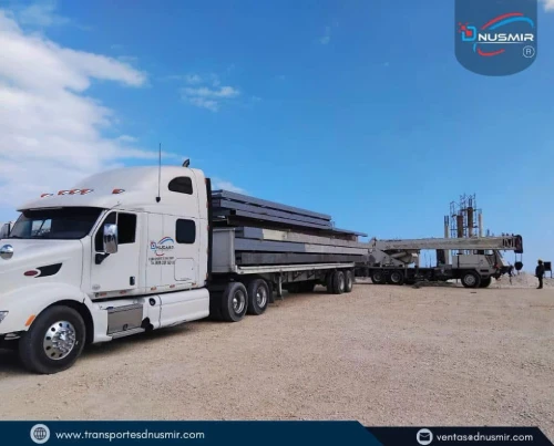 Camión de plataforma transportando vigas estructurales de gran tamaño en zona de obra